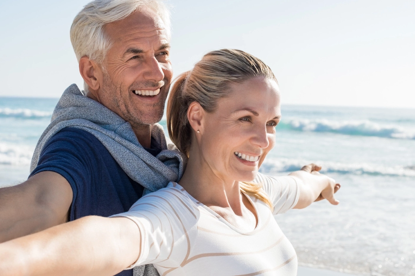 Happy couple at the beach, arms outstretched, smiling, sunny day, sea in background.